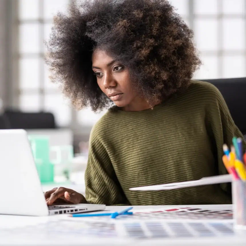 A Black female designer sat at her desk comparing some colours on a print out against her computer screen.