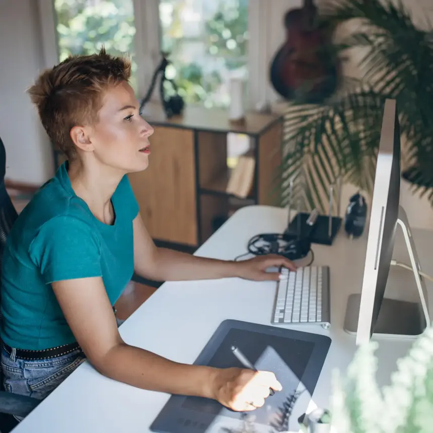 White female designer with short hair sitting at a desk in a modern working environment using a digital drawing pad while concentrating on her main screen.