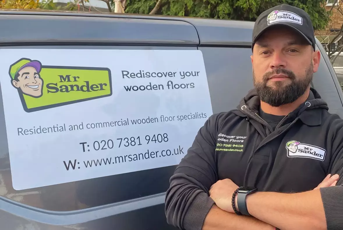 A Mr Sander employee stood next to a van with a Mr Sander magnetic sign on. Employee is wearing branded work clothing showing a the full colour Mr Sander logo on a black polo shirt and baseball cap