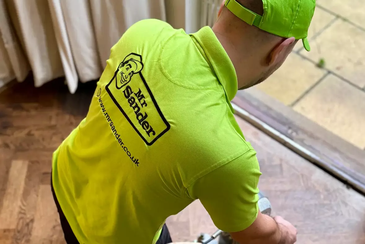 An employee sanding a wooden floor. Employee is wearing branded work clothing showing a solid black logo on a green polo shirt.